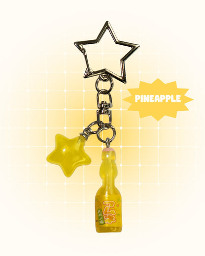 Keychain with star-shaped charm and pineapple soda bottle charm on a yellow gradient background